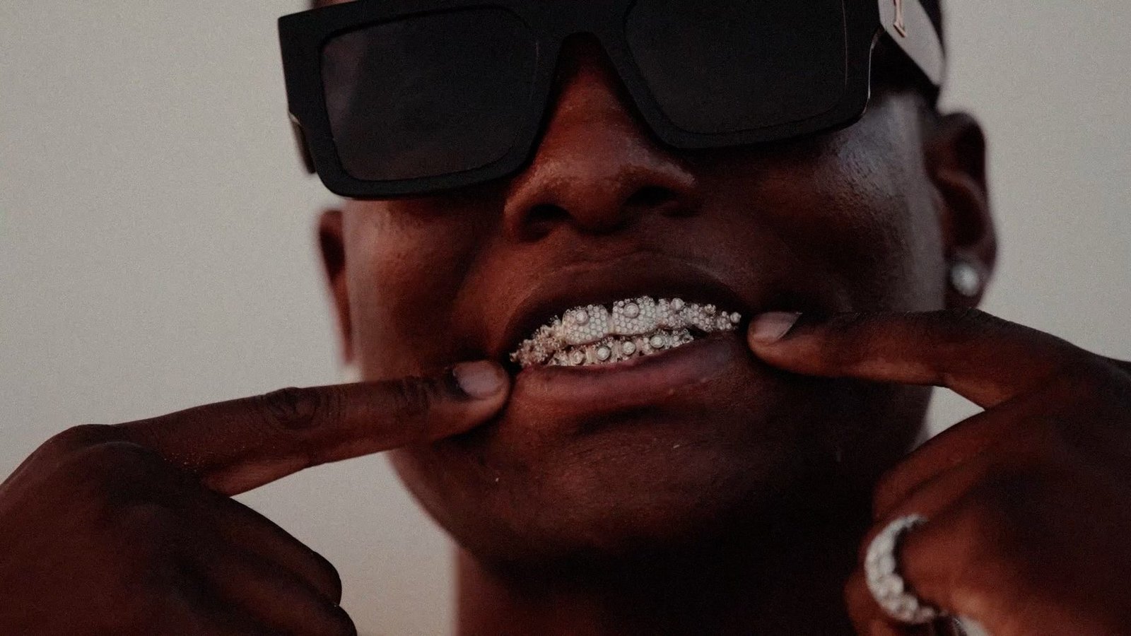 Music video still: a fashion-forward artist in bold sunglasses and a jewelled grill, warm cinematic tones.